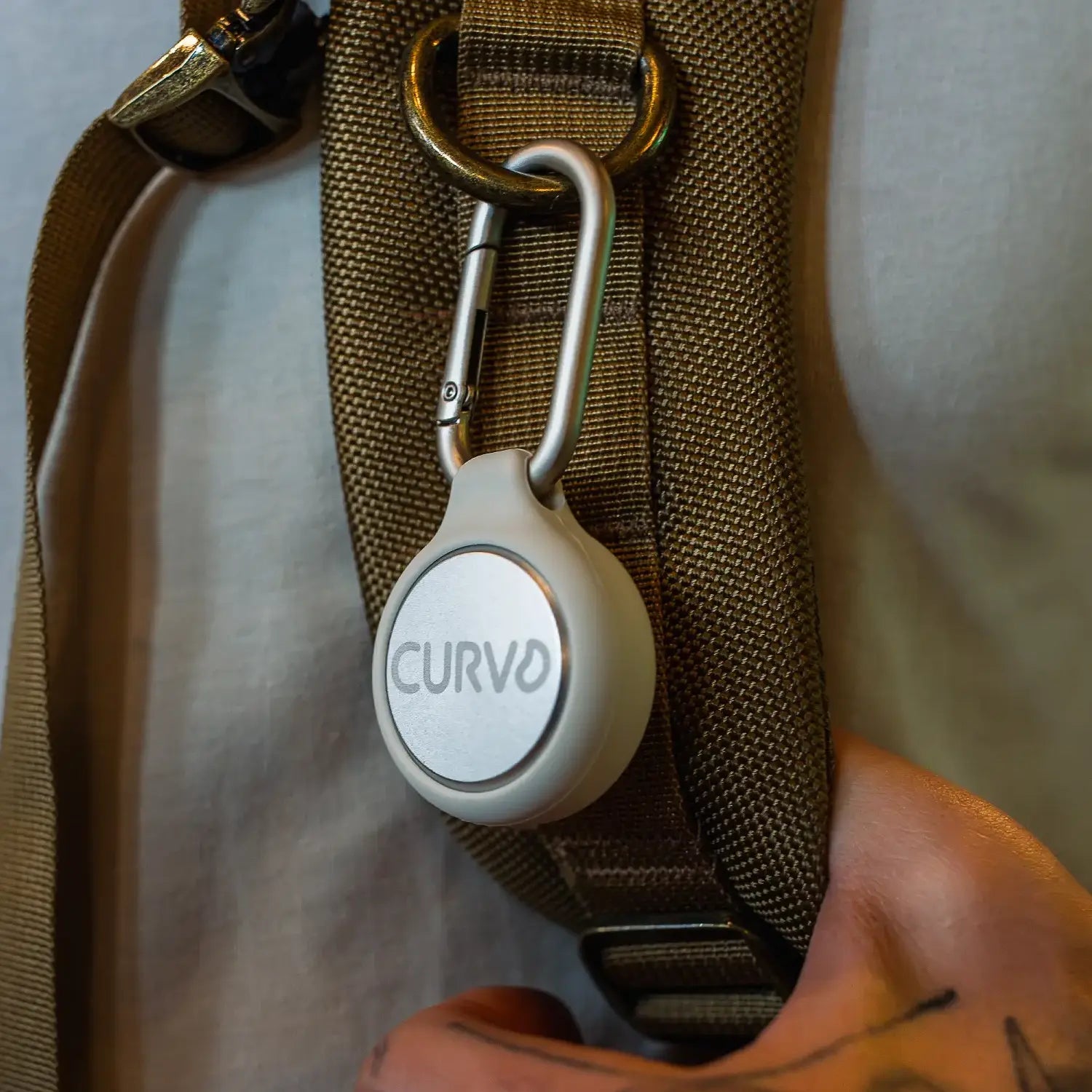 A circular white device labeled "CURVD" is clipped to a brown strap using a metallic carabiner. The strap, which appears to be part of a backpack against a white shirt, securely holds this **Premium Carrying Case Plus Clip by CURVD Earplugs**. Text at the bottom reads, "CLIPS ANYWHERE.