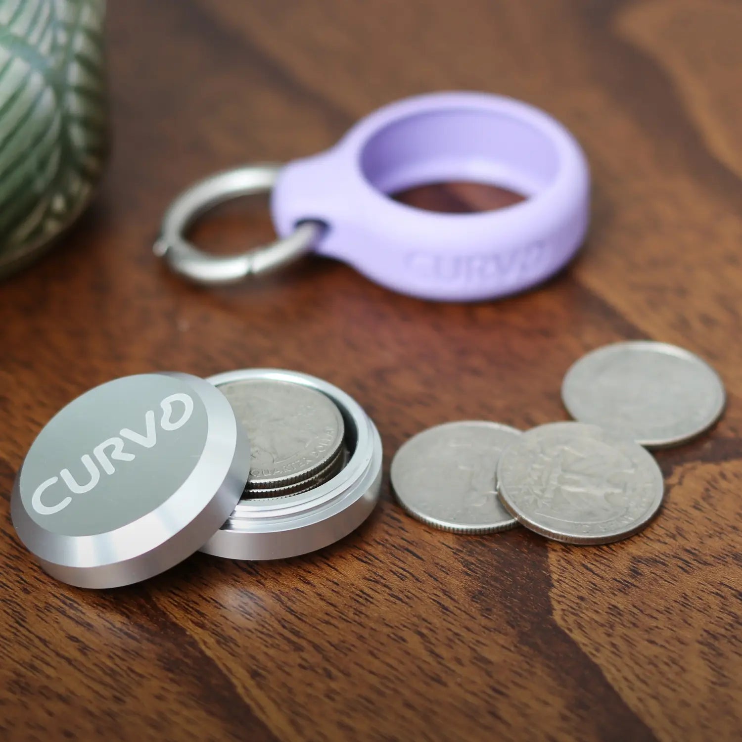 A small round metal container with the word "CURV3" on the lid is open, revealing coins inside. Next to it are a few coins and a purple ring with "CURV3" inscribed, all on a wooden surface. The **Premium Carrying Case Plus Clip** sits beside them. The text **"TINY TREASURES"** from **CURVD Earplugs** is at the bottom of the image.