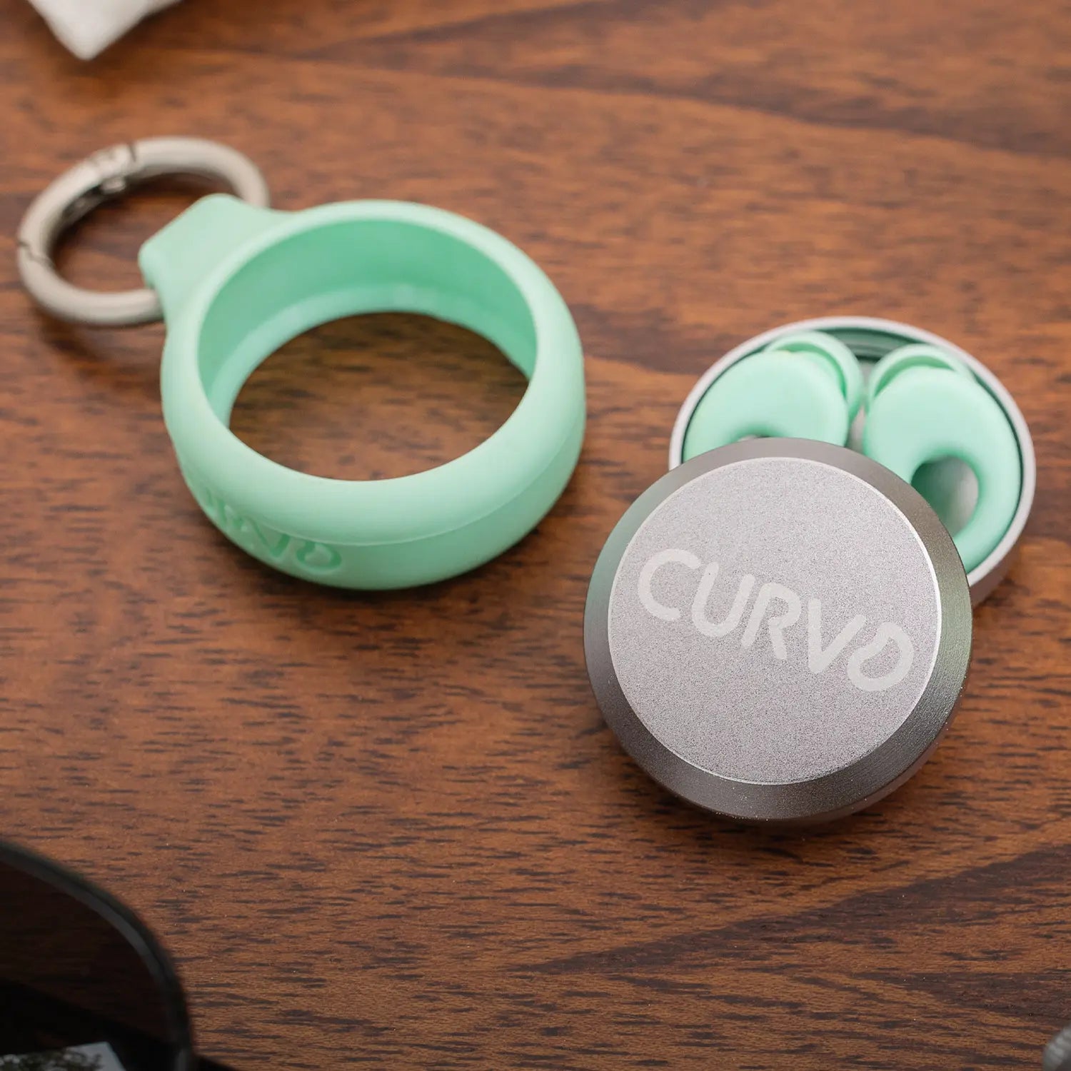 A set of CURVD Earplugs lies on a wooden surface. The CURVD Earplugs are placed in an open, round, silver Premium Carrying Case Plus Clip with the brand name "CURVD" displayed on the lid. Beside the case is a green silicone keychain clip holder. The word "EARPLUGS" is at the bottom of the image.