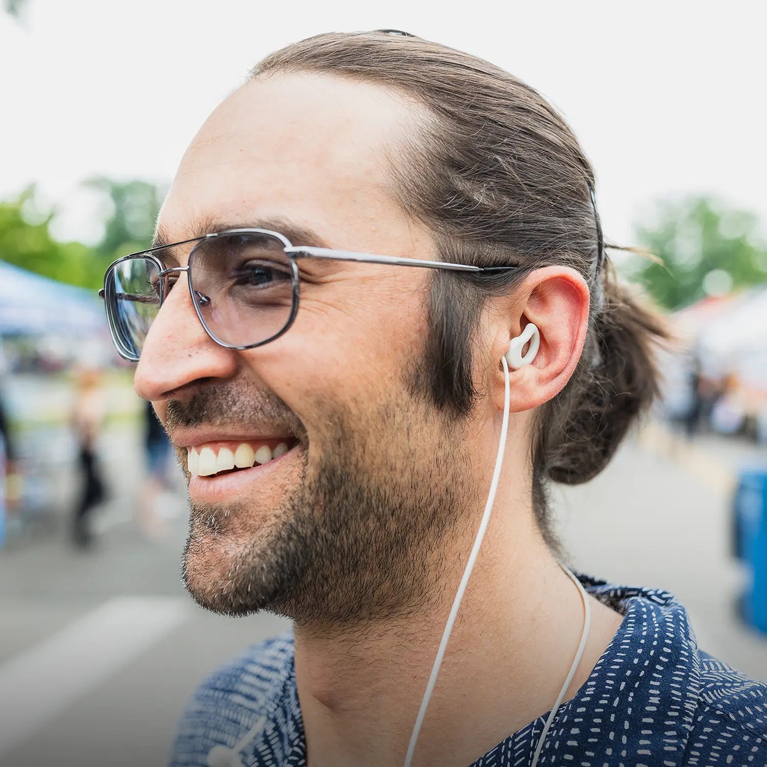 Man wearing CURVD earplugs with a honey-colored lanyard at an outdoor event, smiling and enjoying his surroundings.