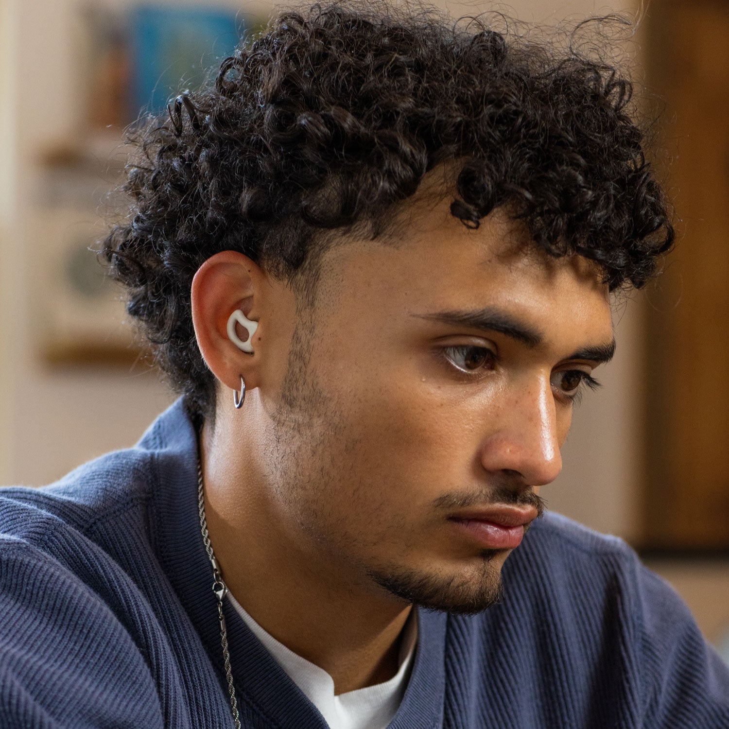 A person with curly hair and a short beard is wearing a single CURVD Earplugs Everyday Earplugs + Case Clip. They appear focused and are looking down. The background is blurred, highlighting the person's concentration. The text "ENHANCED FOCUS" is displayed at the bottom of the image.