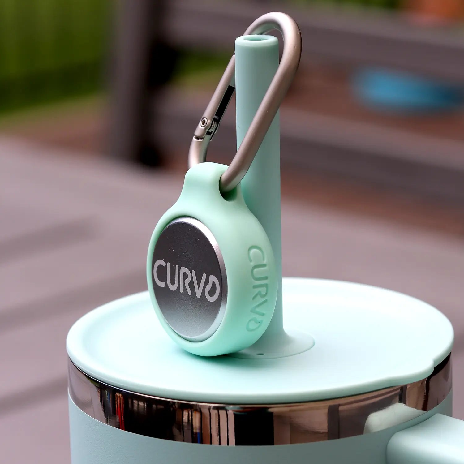 A teal silicone holder with a metal round button labeled "CURVD" and a carabiner clip is attached to the lid of a cup. The background is blurred. The text "SLEEK AND STYLISH" is displayed at the bottom of the image, hinting at CURVD Earplugs' Everyday Earplugs + Case Clip designed for sound reduction.