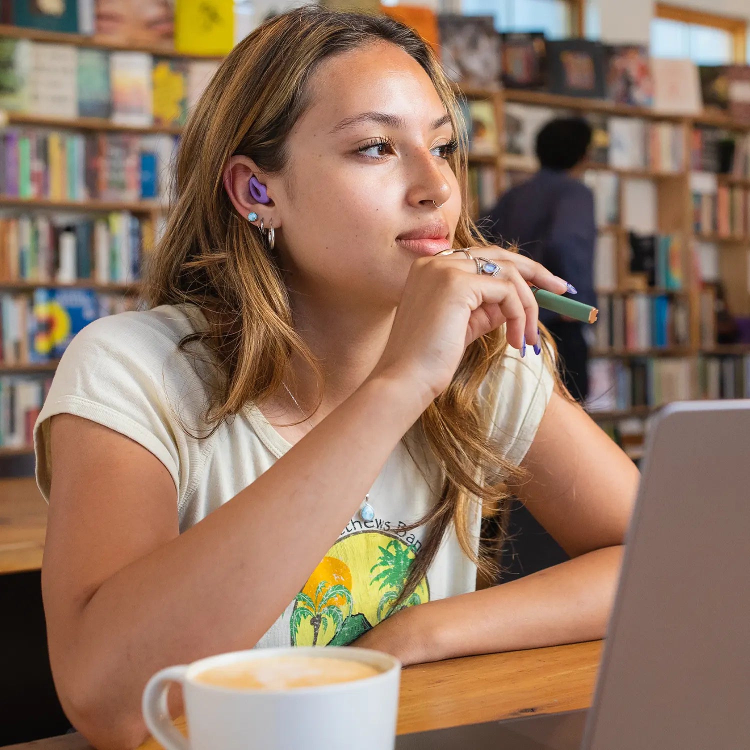 Young woman sitting in a library with purple CURVD Earplugs, looking thoughtfully at her laptop. She holds a pen in one hand, appearing focused and engaged in her work.