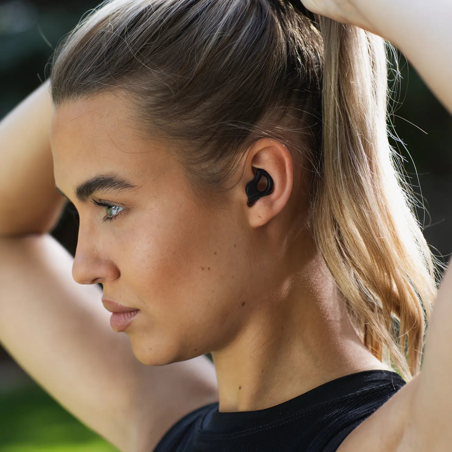 A person with long blonde hair is adjusting their ponytail while wearing a black earbud. They have a focused expression, and the text "UMATCHED COMFORT" is overlaid at the bottom of the image, highlighting the CURVD Earplugs Everyday Earplugs + Case Clip known for their excellent sound reduction.